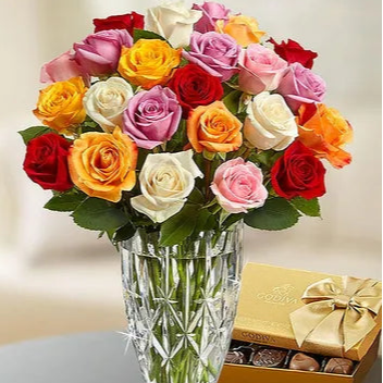 Bouquet of colorful roses in a clear vase with a gold box of chocolates on a reflective surface.